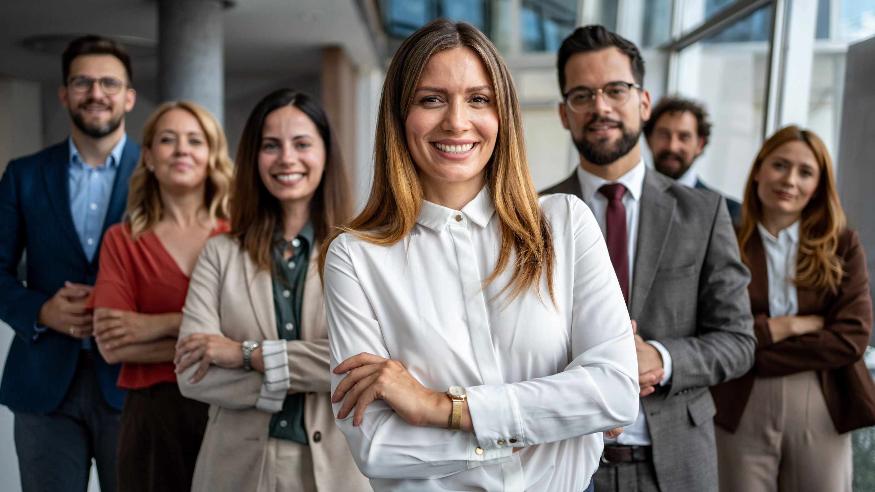 Female leader smiling with crossed arms, standing confidently in front of her diverse business team in a modern office setting, representing teamwork and success
